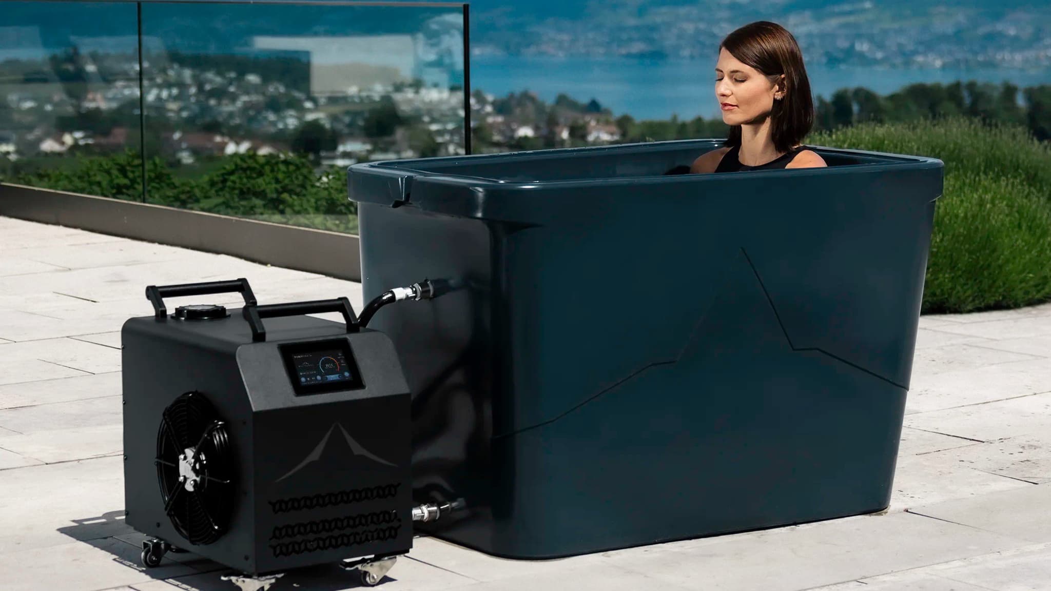 A woman in a Theralpine Rhone ice bath on a sunny terrace above a lake, with the Chiller Pro connected beside the tub.